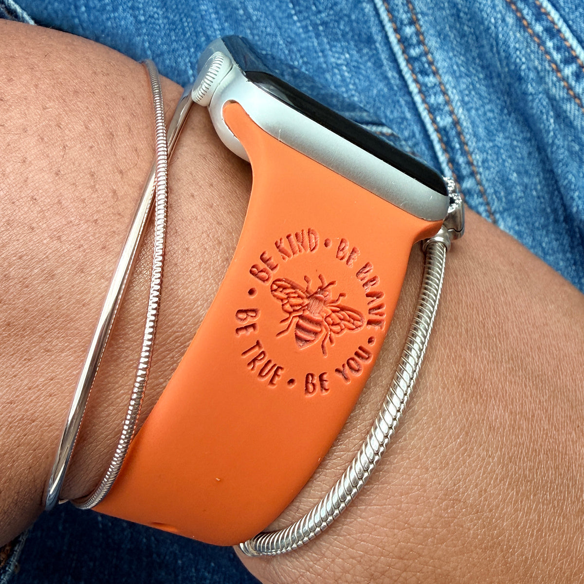 Rust Brave watch band for Apple Watch engraved with the Be Kind Be Brave Bee design, worn on a wrist with silver bracelets