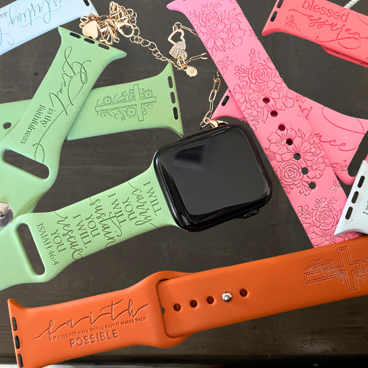 Flat lay of faith engraved watch band for Apple Watchs in green, pink and rust on dark wood