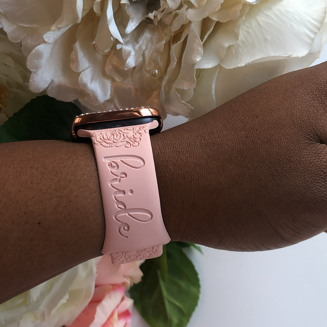 Blush floral watch band for Apple Watch engraved with the word bride, worn on a wrist beside ivory flowers